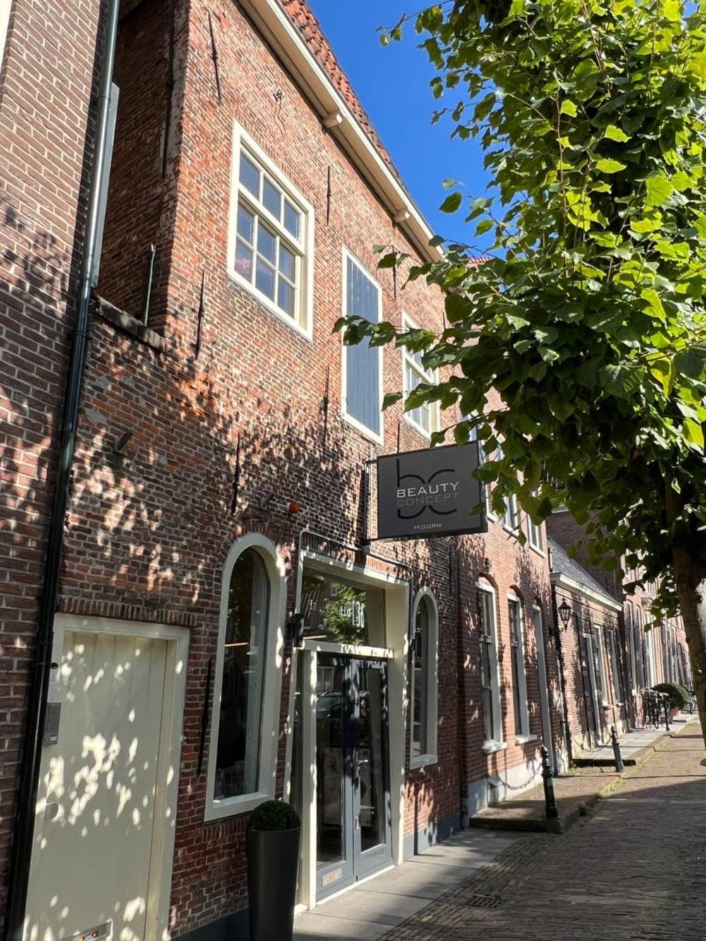 Het pand van Beauty Concept Hoorn aan de Ramen 36: een oud bakstenen gebouw met boogramen en een uithangbord, aan een straat met bomen